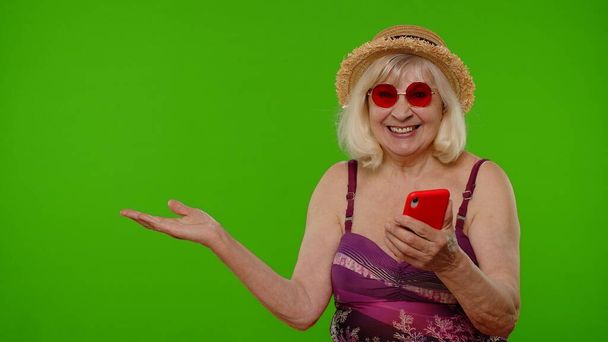 Senior nyugdíjas nő turista fürdőruha pont kézzel mutatja hirdetési tartalom mobiltelefon - Fotó, kép