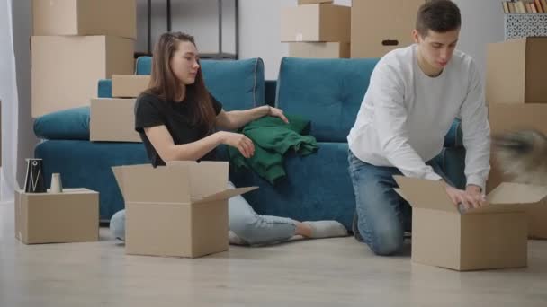 La pareja feliz está empacando cosas en cajas de cartón para mudarse a una casa nueva. El perro pasea por la habitación y observa. - Metraje, vídeo