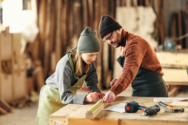 Положительная молодая ремесленница делает отметки на деревянных деталях, работая с партнером-мужчиной, работая вместе в столярном цехе. - Фото, изображение