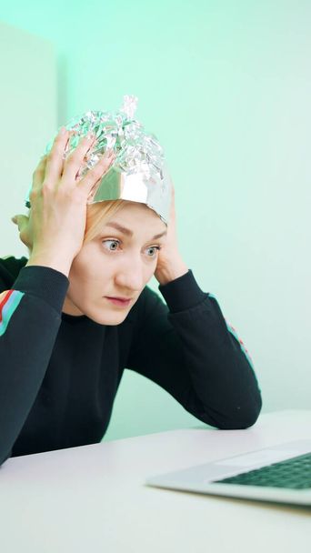 Vertikal. Ängstliche junge Frau mit Aluminiumhut surft in den sozialen Medien. Verschwörungstheorie über 5g Mikrowellen  - Foto, Bild