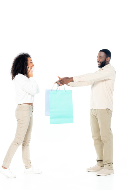 sonriente afroamericano hombre dando bolsas de compras a la esposa sorprendida en blanco - Foto, Imagen