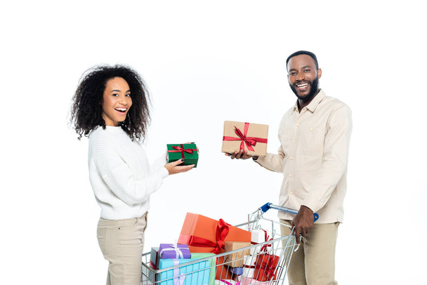vidám afro-amerikai pár néz kamera, miközben tartja ajándékdobozok közelében bevásárlókocsi elszigetelt fehér - Fotó, kép