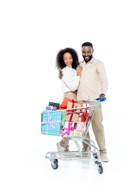 veselý africký americký pár objímající v blízkosti nákupního košíku s dárky krabice na bílém - Fotografie, Obrázek