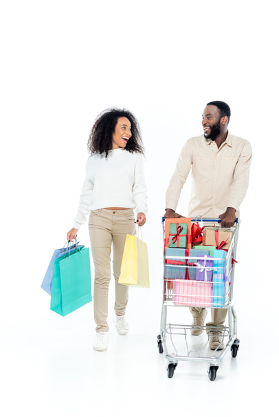 laughing african american couple looking at each other while walking with purchases on white - Photo, Image