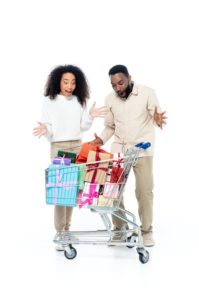 užaslý africký americký pár ukazuje wow gesto v blízkosti nákupní vozík plný dárků na bílém - Fotografie, Obrázek