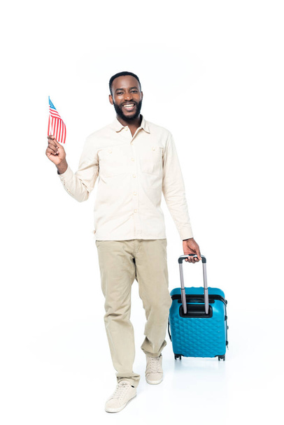 homme afro-américain joyeux tenant petit drapeau des Etats-Unis tout en marchant avec une valise sur blanc - Photo, image