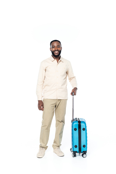 glücklich afrikanisch-amerikanischer Mann blickt in die Kamera, während er mit Reisetasche auf weiß steht - Foto, Bild
