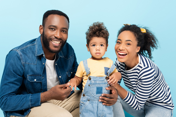 alegre africano americano pareja con pequeño hijo sonriendo a cámara aislado en azul - Foto, imagen