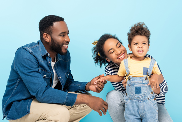 smiling african american man holding hand of cheerful son near happy wife isolated on blue - Photo, Image