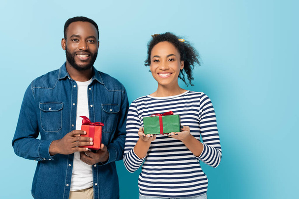 feliz pareja afroamericana sosteniendo cajas de regalo mientras mira la cámara en azul - Foto, Imagen