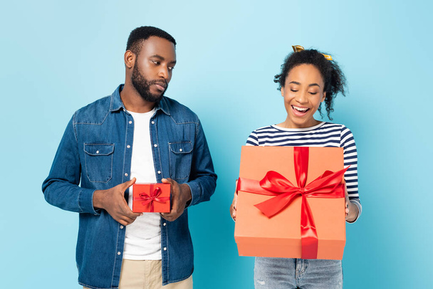 beleidigt afrikanisch-amerikanischer Mann mit kleinem Geschenkkarton schaut erstaunt Ehefrau mit großem Geschenk auf blau an - Foto, Bild