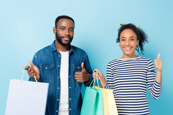 fröhliches afrikanisch-amerikanisches Paar hält Einkaufstüten in der Hand und zeigt Daumen hoch auf blau - Foto, Bild