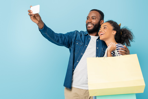 heureux homme afro-américain prenant selfie avec femme gaie tenant des sacs à provisions sur bleu - Photo, image