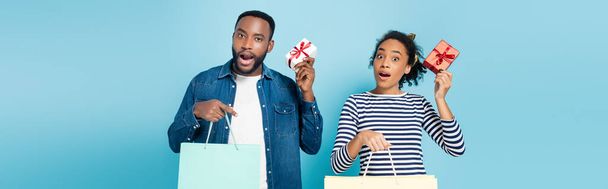 amazed african american couple holding gift boxes and shopping bags on blue, banner - Photo, Image