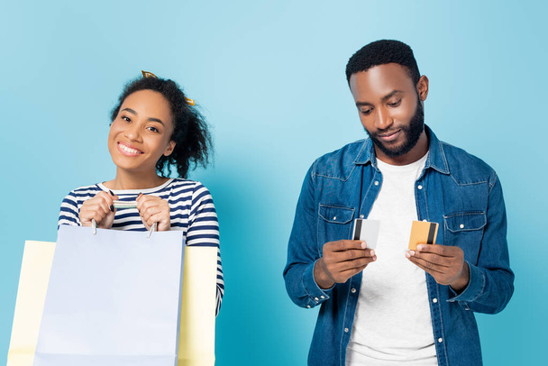 Afro-Amerikalı bir adam elinde kredi kartları, mutlu karısının yanında mavi torbalarla. - Fotoğraf, Görsel