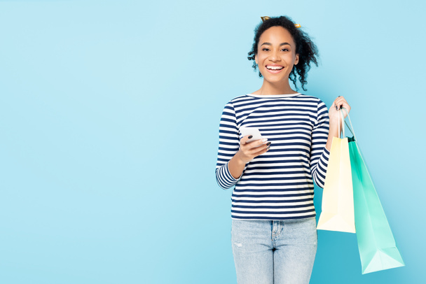 alegre mujer afroamericana con teléfono móvil y bolsas de compras mirando a la cámara aislada en azul - Foto, imagen