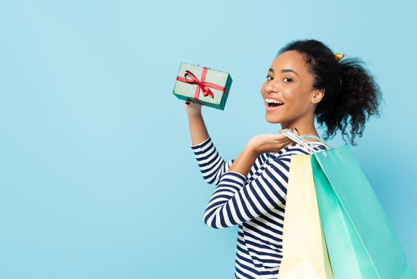 mujer afroamericana asombrada mostrando presente mientras sostiene bolsas de compras aisladas en azul - Foto, Imagen