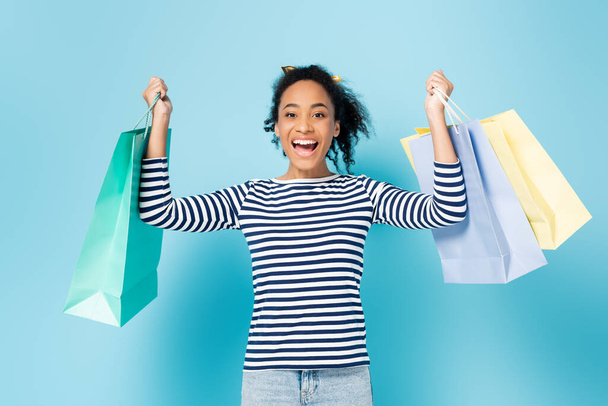 mulher americana africana surpreendido olhando para a câmera enquanto segurando sacos de compras em azul - Foto, Imagem