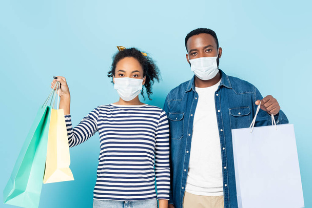 jong Afrikaans amerikaans paar in medische maskers houden boodschappentassen op blauw - Foto, afbeelding