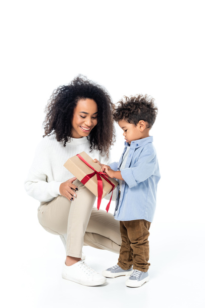 Αφρικανός Αμερικανός παιδί άνοιγμα κουτί δώρου κοντά χαμογελαστή μητέρα σε λευκό - Φωτογραφία, εικόνα