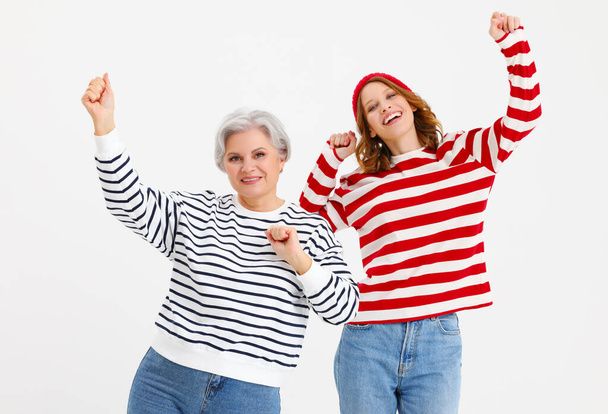Pozytywny w średnim wieku siwe włosy kobieta i nastoletnia córka w kolorowe paski koszule i dżinsy zabawy i tańca z ramionami podniesione na białym tle - Zdjęcie, obraz