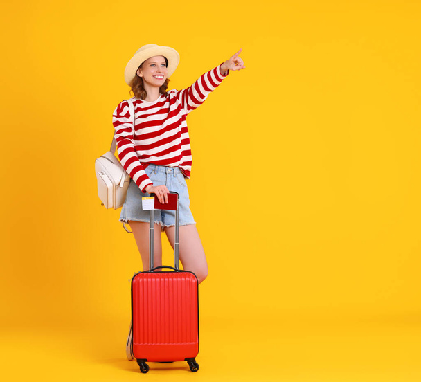 Uzun boylu, rahat kıyafetli ve şapkalı genç bir bayan turist sarı duvarın yanında durup tatil sırasında uçmayı beklerken bavulunu gösteriyordu. - Fotoğraf, Görsel