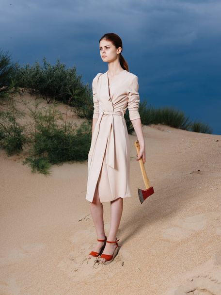 Fashionable woman in a sundress with an ax in her hands and in red shoes stands on the sand on the beach - Photo, image