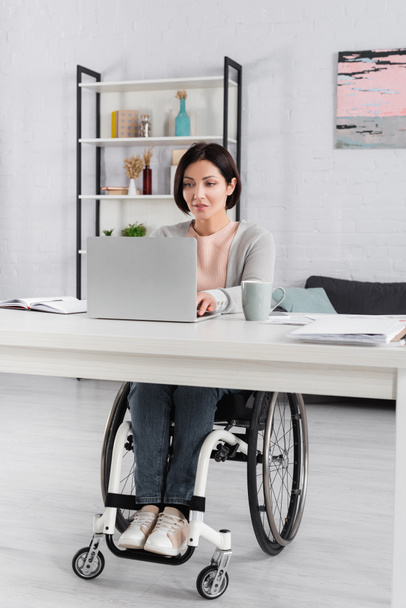 Fincanın yanında dizüstü bilgisayar kullanan engelli kadın ve masadaki bulanık ön planda belgeler  - Fotoğraf, Görsel