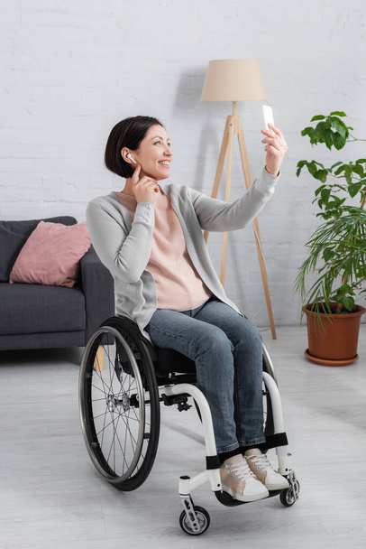 Mujer discapacitada feliz en auriculares usando teléfono inteligente en casa  - Foto, Imagen
