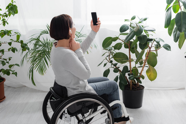 Frau im Rollstuhl benutzt Kopfhörer und Smartphone zu Hause  - Foto, Bild