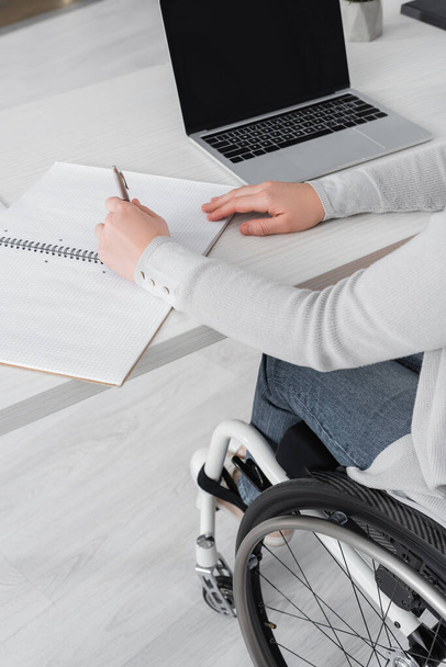 Ausgeschnittene Ansicht einer Frau im Rollstuhl, die auf einem Notizbuch neben einem Laptop mit leerem Bildschirm auf dem Tisch schreibt  - Foto, Bild