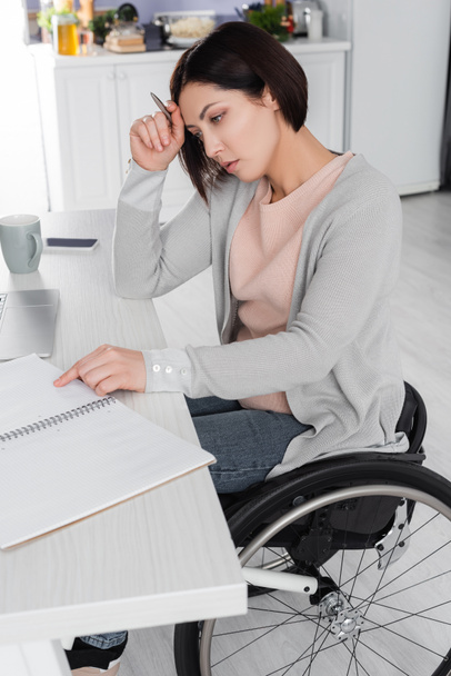 Ελεύθερος επαγγελματίας σε αναπηρική καρέκλα κρατώντας στυλό κοντά σημειωματάριο και gadgets στο τραπέζι  - Φωτογραφία, εικόνα