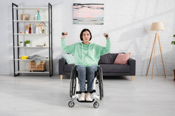 Μελαχρινή γυναίκα που γυμνάζεται με αλτήρες σε αναπηρική καρέκλα και κοιτάζοντας την κάμερα στο σπίτι   - Φωτογραφία, εικόνα