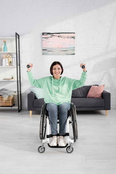 Mujer alegre en silla de ruedas mirando a la cámara mientras entrena con pesas  - Foto, imagen