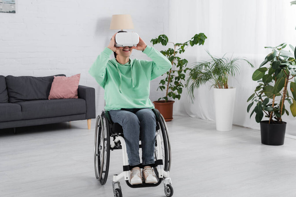 Χαρούμενη γυναίκα σε αναπηρική καρέκλα χρησιμοποιώντας ακουστικά vr στο σαλόνι  - Φωτογραφία, εικόνα