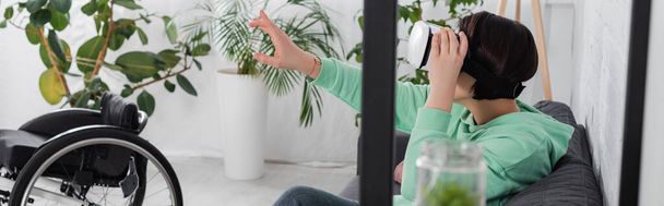 Brunette nainen käyttää virtuaalitodellisuus kuulokkeet lähellä pyörätuolia kotona, banneri  - Valokuva, kuva