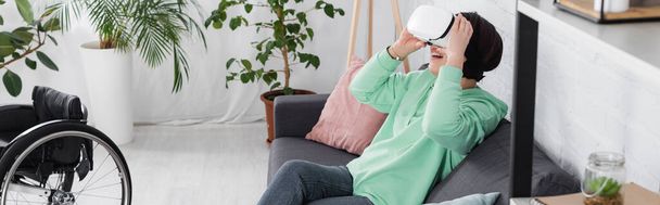 Positiivinen nainen vr kuulokkeet istuu sohvalla lähellä pyörätuolia olohuoneessa, banneri  - Valokuva, kuva