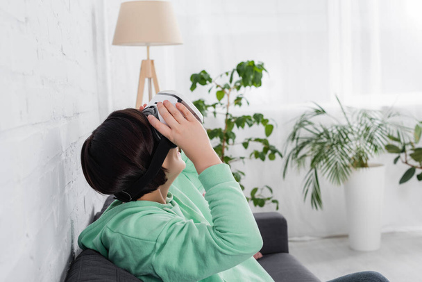 Morena mulher segurando vr fone de ouvido no sofá na sala de estar  - Foto, Imagem