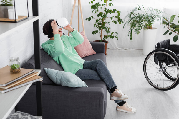 Mujer feliz disgustado en auriculares vr sentado en el sofá cerca de la silla de ruedas y armario en primer plano borrosa  - Foto, Imagen