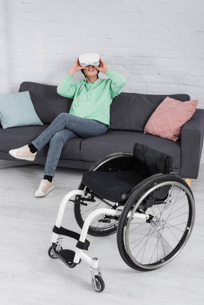 Uśmiechnięta niepełnosprawna kobieta w zestawie słuchawkowym VR siedzi na kanapie obok wózka inwalidzkiego  - Zdjęcie, obraz