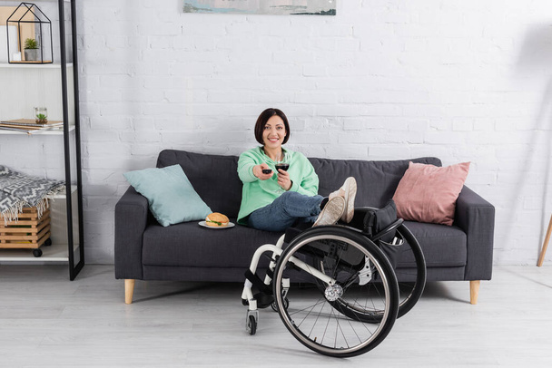 Lächelnde Frau mit einem Glas Wein vor dem Fernseher in der Nähe von Burger und Rollstuhl im Wohnzimmer  - Foto, Bild