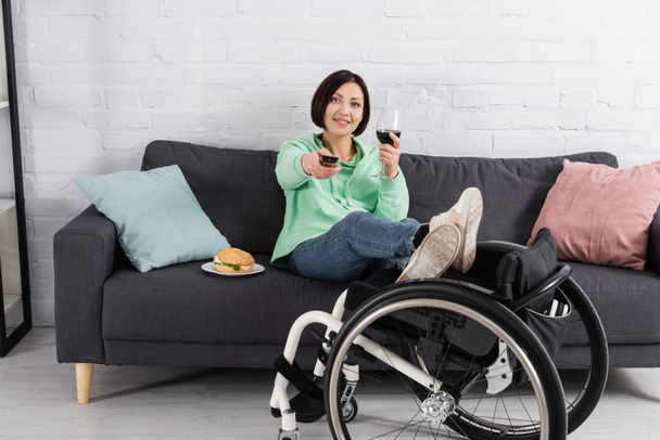 Gehbehinderte Frau mit Fernbedienung und Glas Wein sitzt in der Nähe von Hamburger und Rollstuhl zu Hause  - Foto, Bild