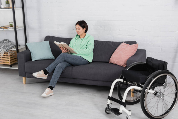 Brunette femme livre de lecture sur canapé près de fauteuil roulant dans le salon  - Photo, image