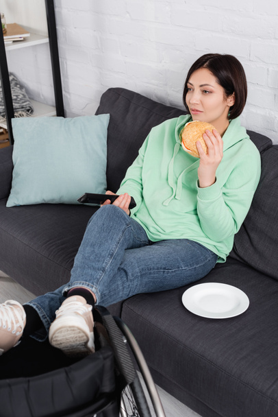Žena s hamburgerem drží dálkové ovládání v blízkosti invalidního vozíku doma  - Fotografie, Obrázek