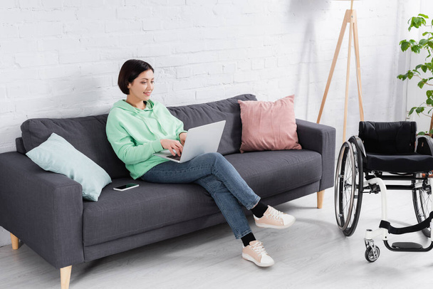 Улыбающаяся женщина с ноутбука рядом со смартфоном и инвалидной коляской в гостиной  - Фото, изображение