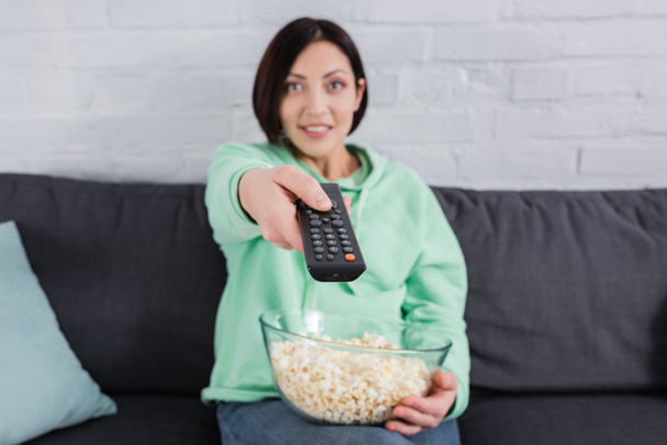 Пульт дистанционного управления в руке улыбающейся женщины с попкорном на диване на размытом фоне  - Фото, изображение