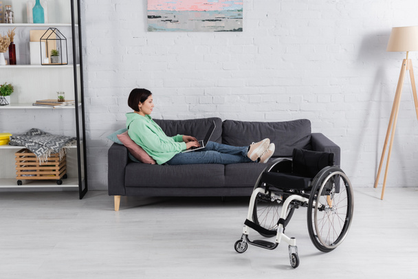 Πλευρική άποψη της δυσαρεστημένης γυναίκας με τη χρήση φορητού υπολογιστή στον καναπέ κοντά σε αναπηρικό καροτσάκι  - Φωτογραφία, εικόνα