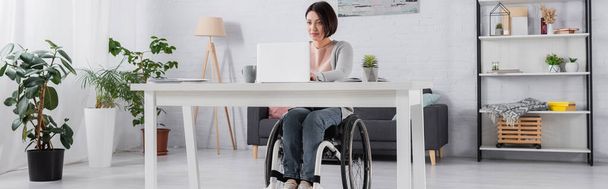 Wolny strzelec na wózku inwalidzkim przy użyciu laptopa w pobliżu kubka w domu, baner  - Zdjęcie, obraz