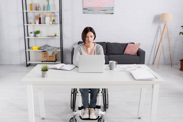 Behinderte Freiberuflerin benutzt Laptop in der Nähe von Dokumenten mit Diagrammen und Tasse auf dem Tisch  - Foto, Bild
