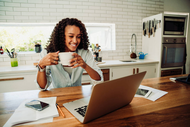 Mezcla de raza sonriente estudiante femenina sosteniendo café caliente sonriendo mientras en videollamada con amigos en el ordenador portátil - Foto, Imagen